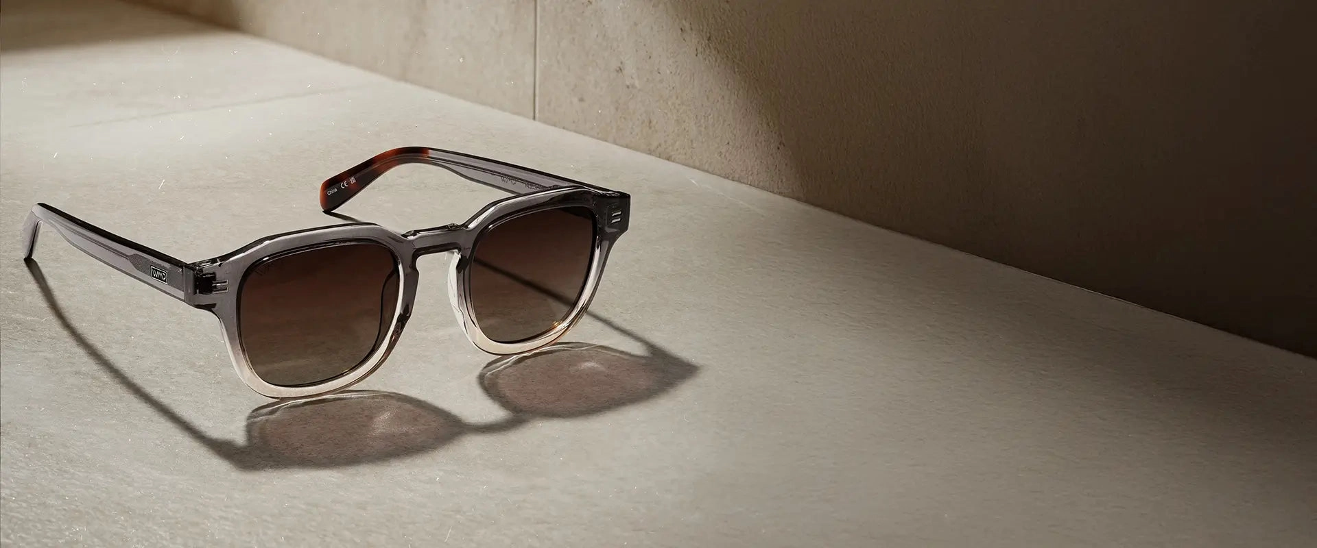 Nelson sunglasses in Graphite Horizon Grey with brown lenses, displayed on a neutral stone surface in warm sunlight.