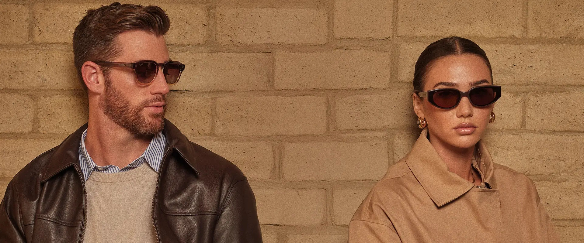 Two models wearing WMP Eyewear sunglasses in warm brown tones, styled in neutral fall layers against a tan brick wall.