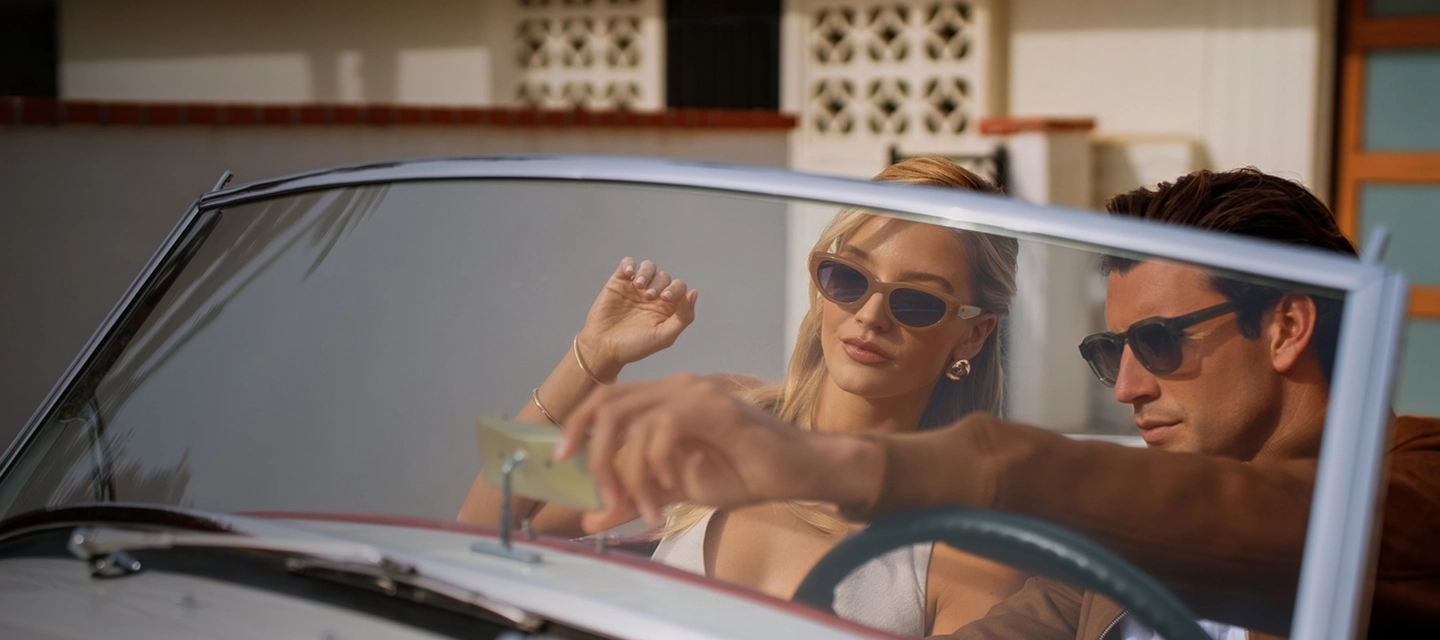 A couple in a convertible in bright sunlight, seen through two sunglass lenses